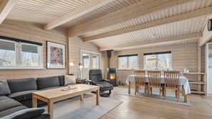L’intérieur chaleureux d’un chalet avec un canapé, une table à manger, des chaises, une cheminée et de grandes fenêtres.
