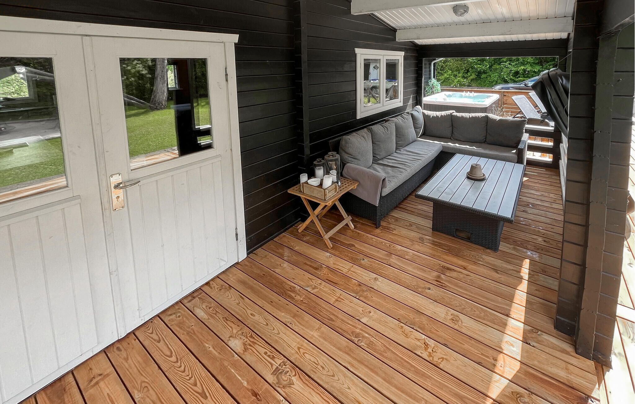 Un espace extérieur abrité doté d’une terrasse en bois, d’un canapé, d’une petite table et d’un jacuzzi visible à travers les fenêtres.