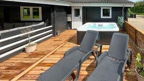 Une terrasse en bois avec deux chaises longues, un jacuzzi et un petit coin salon agrémenté de coussins et d’une plante en pot.