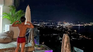 A person in red swim trunks standing on a balcony overlooking a cityscape at night. - Samui Sky Pool Villa (Koh Samui)