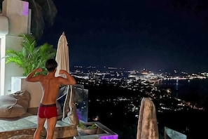 Une personne vêtue d’un maillot de bain rouge, debout sur un balcon surplombant une ville illuminée la nuit.