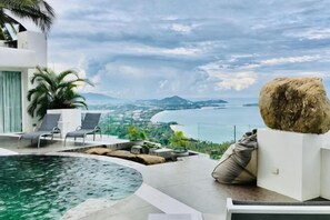 A modern outdoor pool area with lounge chairs, a large rock sculpture, and a scenic view of the sea and mountains.