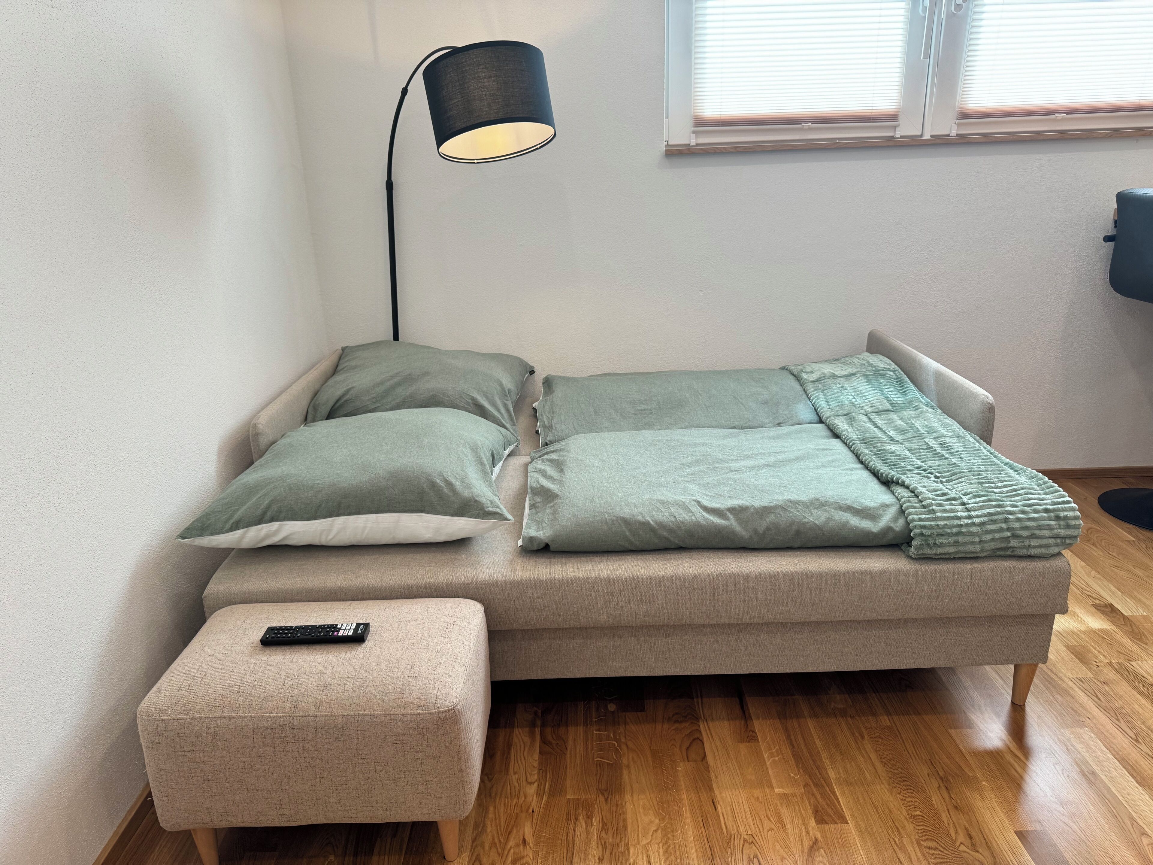 Ein kleiner, gemütlicher Raum mit einem Bett, einem Nachttisch, einer Lampe und einem Fenster mit Jalousien.