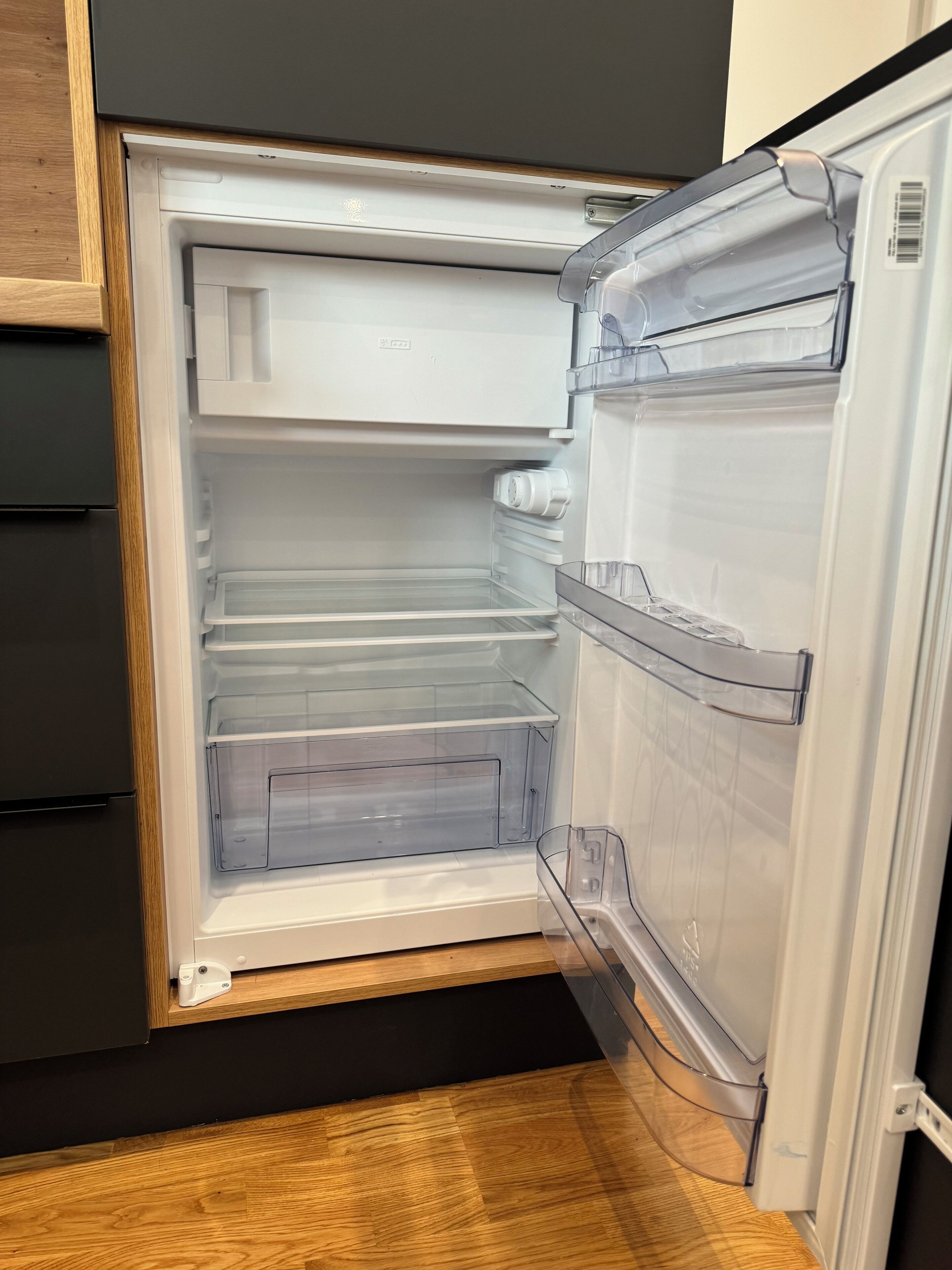 Ett kylskåp med öppna hyllor av genomskinlig plast och en bänkskiva i trä.