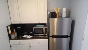 A compact kitchen with white cabinets, a stainless steel refrigerator, a microwave, and a small countertop with a kettle and utensils.