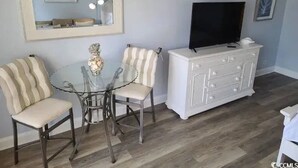 A living room with a round glass table, two striped chairs, a white cabinet with a flat-screen TV, and a framed picture on the wall.