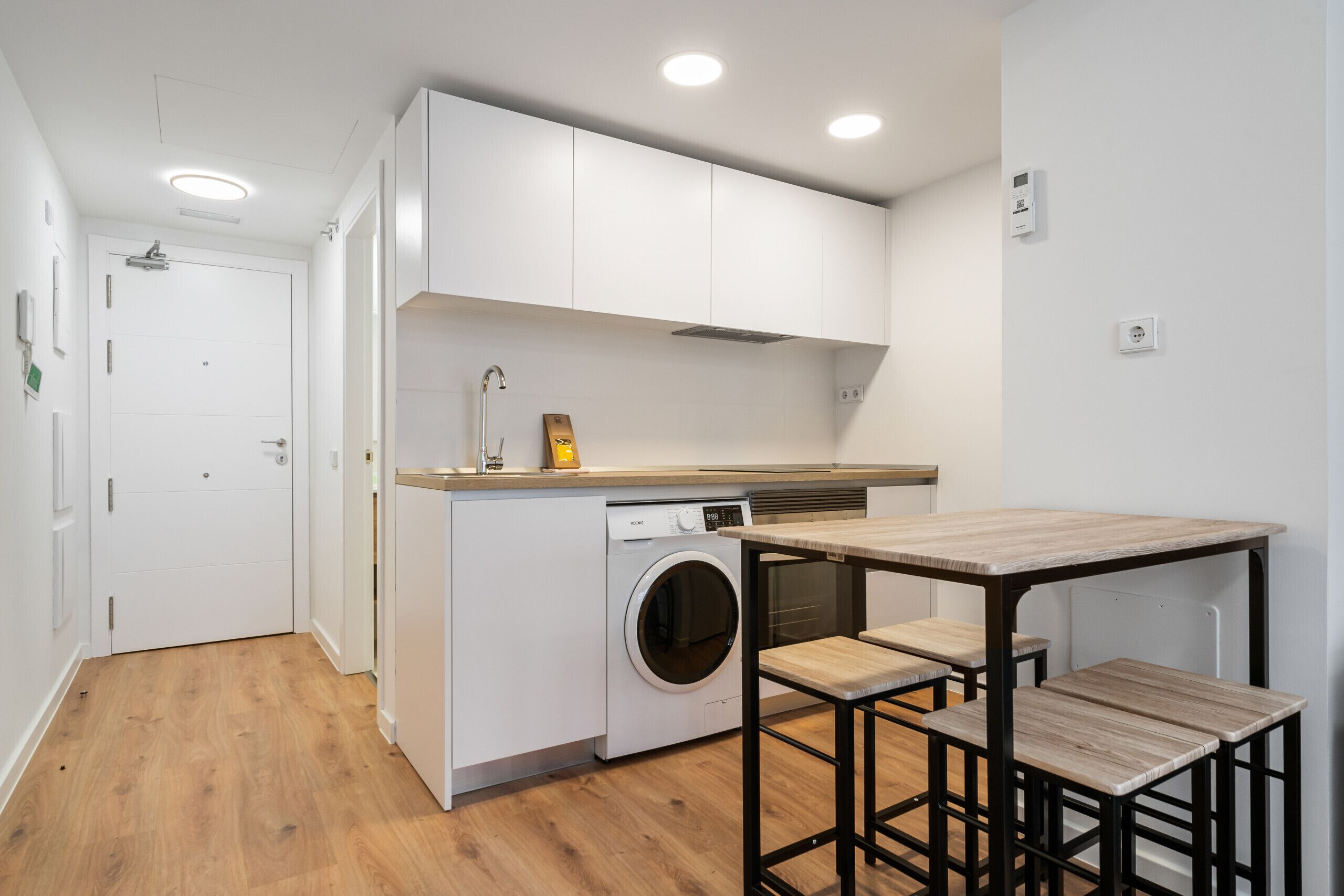 Cocina compacta con lavadora integrada, una mesa de madera y taburetes de bar.