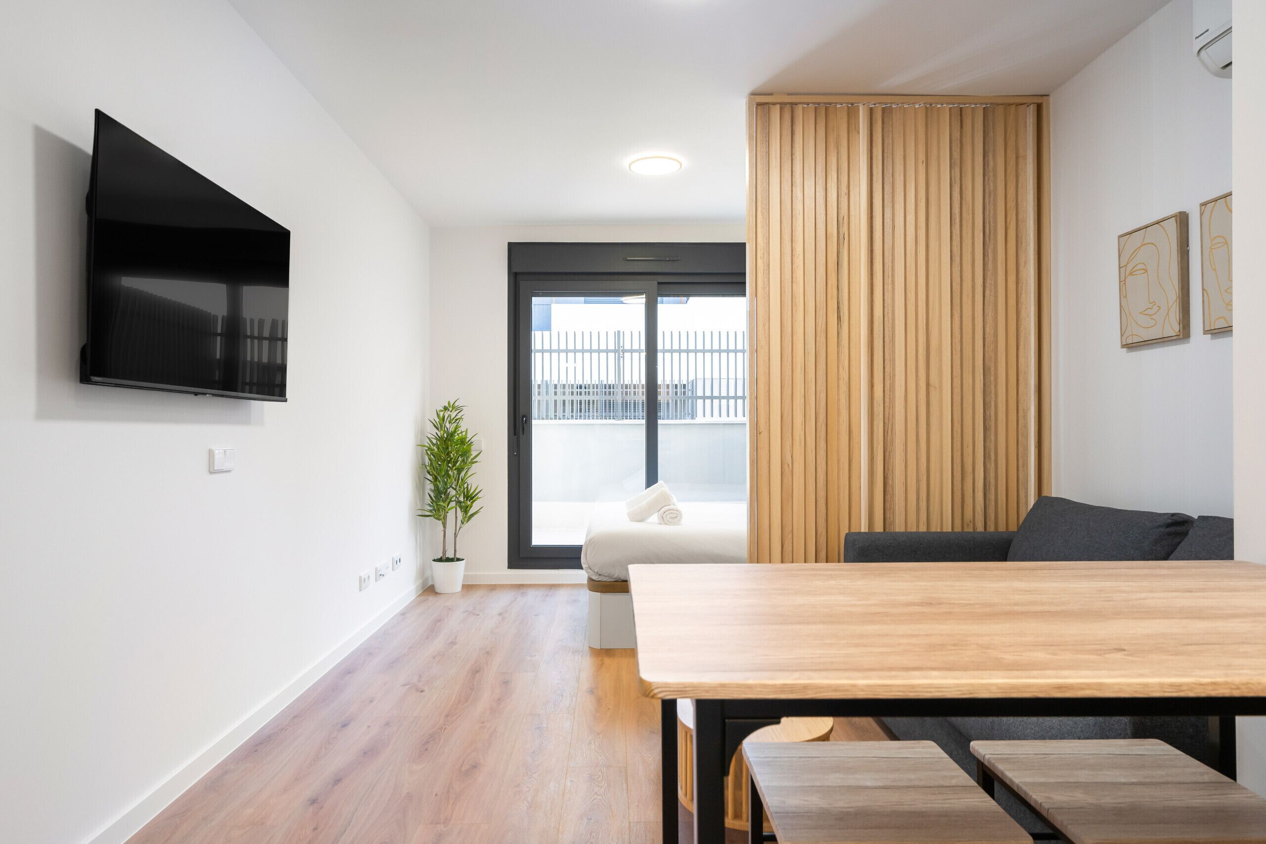 Een moderne woonkamer met een houten eettafel, een flatscreen televisie aan de muur, een groot raam met uitzicht op een omheining en een potplant.