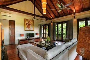 A spacious living room with a large sofa, a coffee table, and a television. There are large windows with curtains, a ceiling fan, and a painting on the wall. - Pulung Villa (Bali)