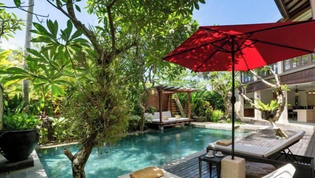 A serene outdoor pool area with lounge chairs and a red umbrella, surrounded by lush greenery. - Pulung Villa (Bali)