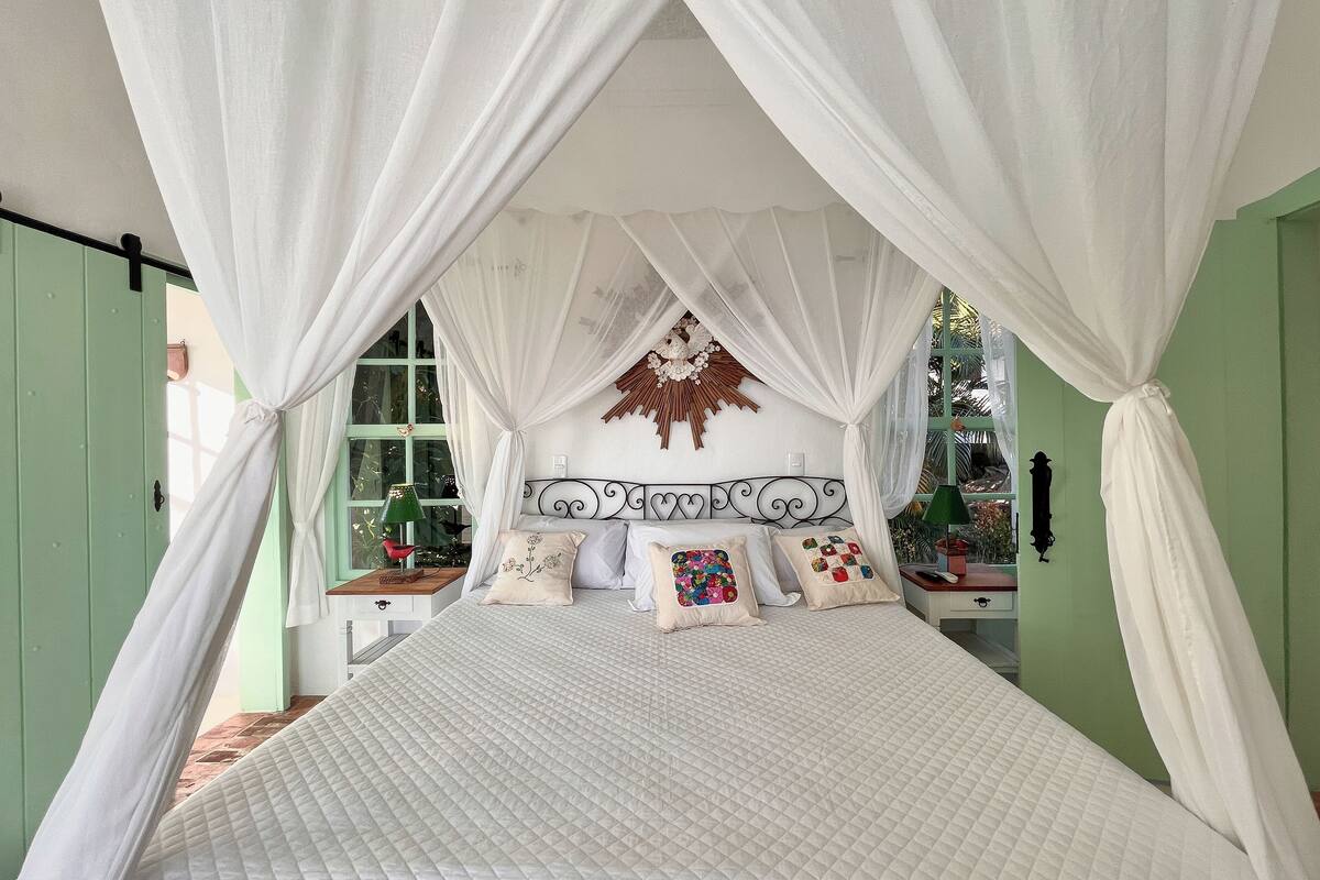 Una cama con colcha blanca acolchada, un tapiz decorativo sobre ella, y una ventana con vistas a la vegetación.