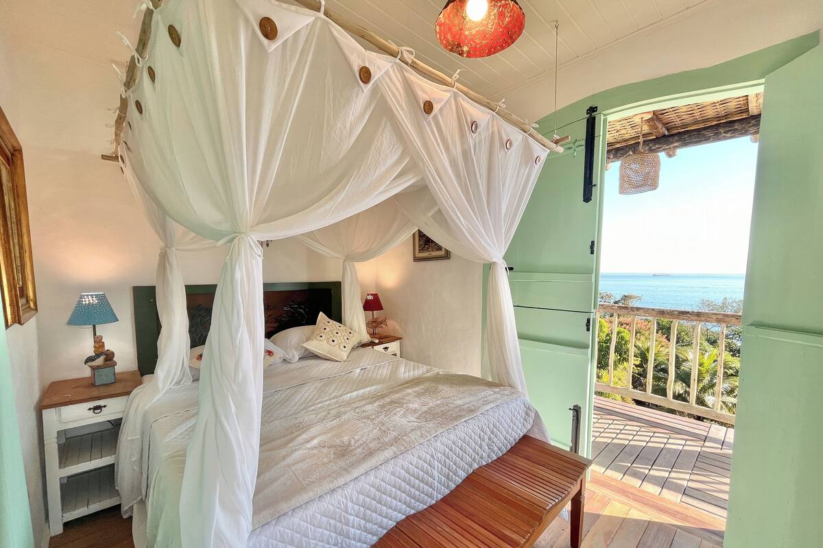 Un dormitorio con cama con dosel, un banco de madera, una mesita de noche con lámpara y vista al mar a través de una puerta abierta.