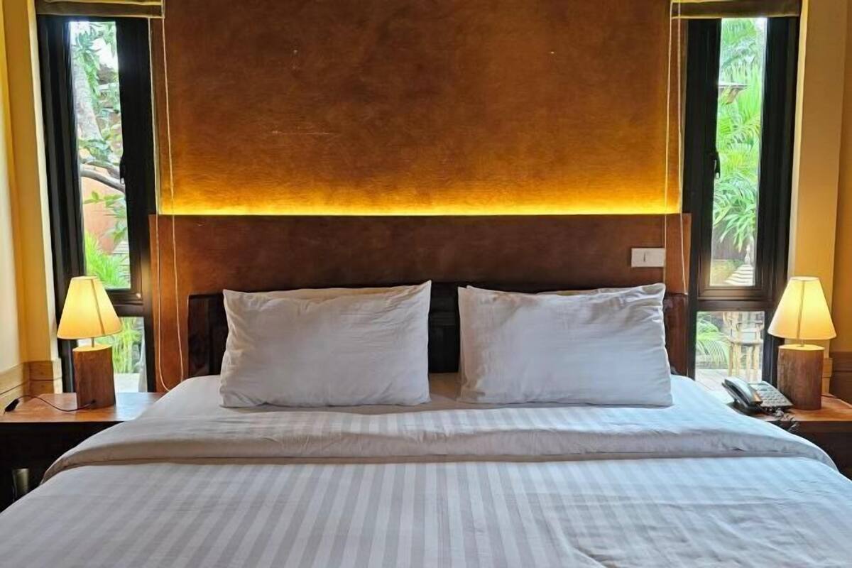 Habitació d'hotel amb un llit gran, dues làmpades de nit i vista a la vegetació per les finestres.