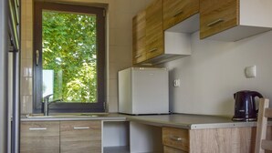 Eine kleine Küche mit Holzschränken, einem weißen Kühlschrank, einem Wasserkocher und einem Fenster mit Blick auf Bäume.
