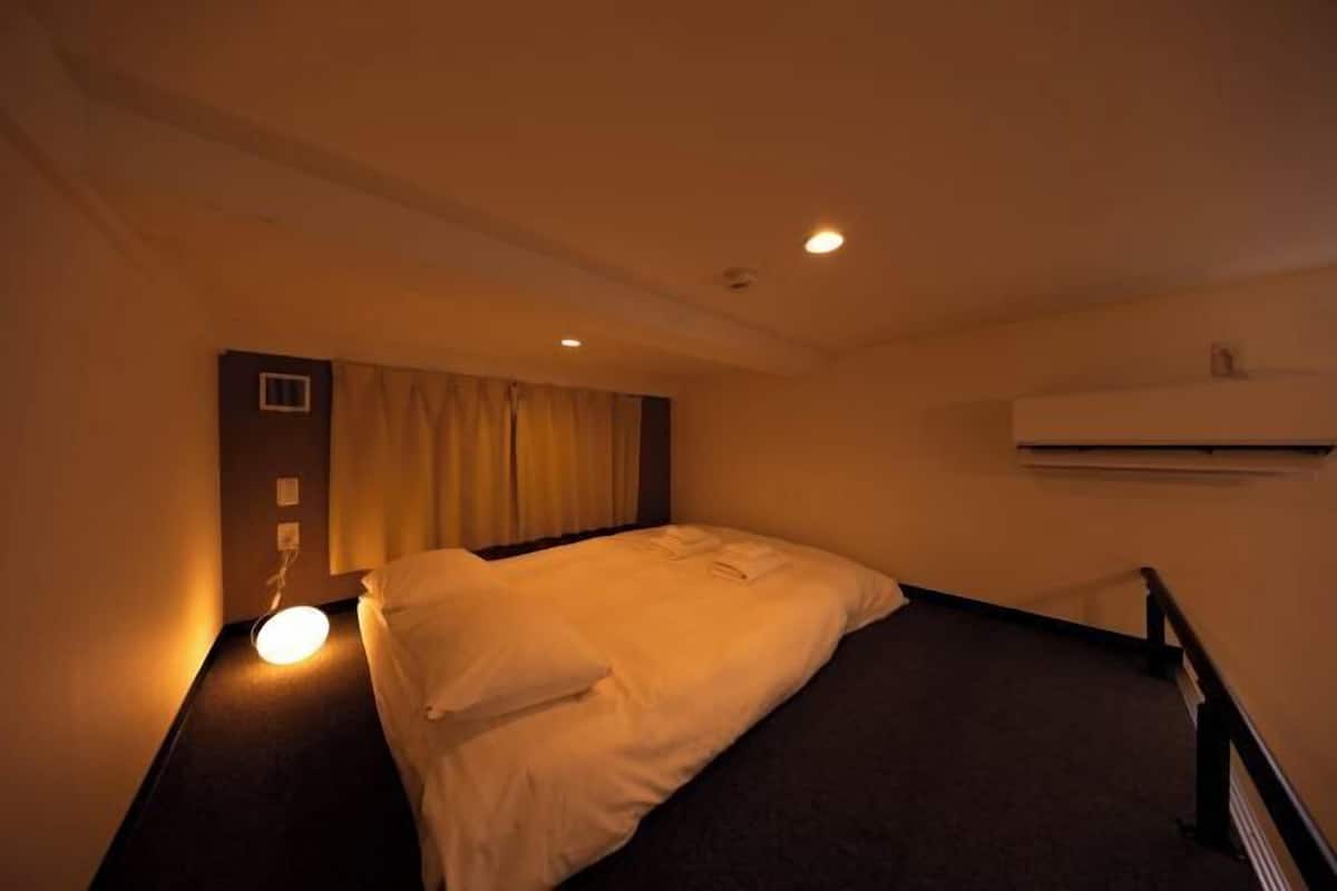 A hotel room with a bed, a wall-mounted air conditioner, and a single light source.