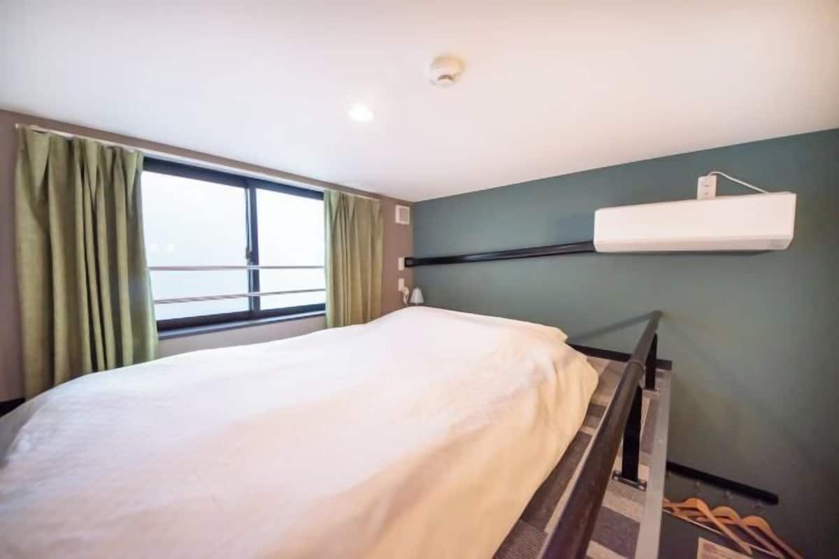 A single bed with a white mattress, a window with curtains, a wall-mounted air conditioning unit, and a small table with a lamp.