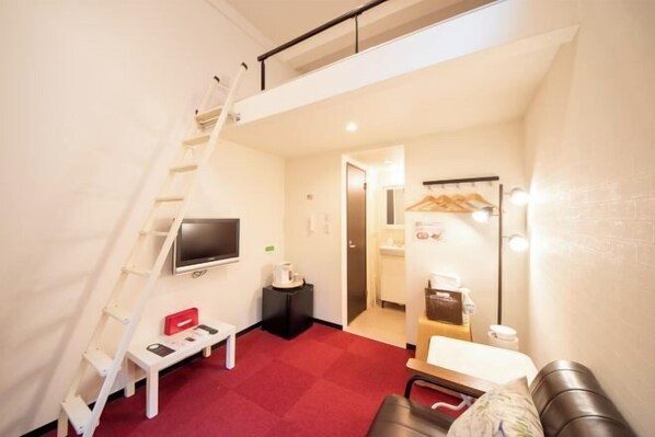 A compact room with a red carpet, a white desk, a black chair, a television, a small table, a ladder, a mirror, and a coffee machine. - Apartment Sanjo (Nara City)