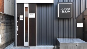 A building entrance with a dark facade, a sign reading 'APARTMENT SANJO,' and a metal structure in front. - Apartment Sanjo (Nara City)