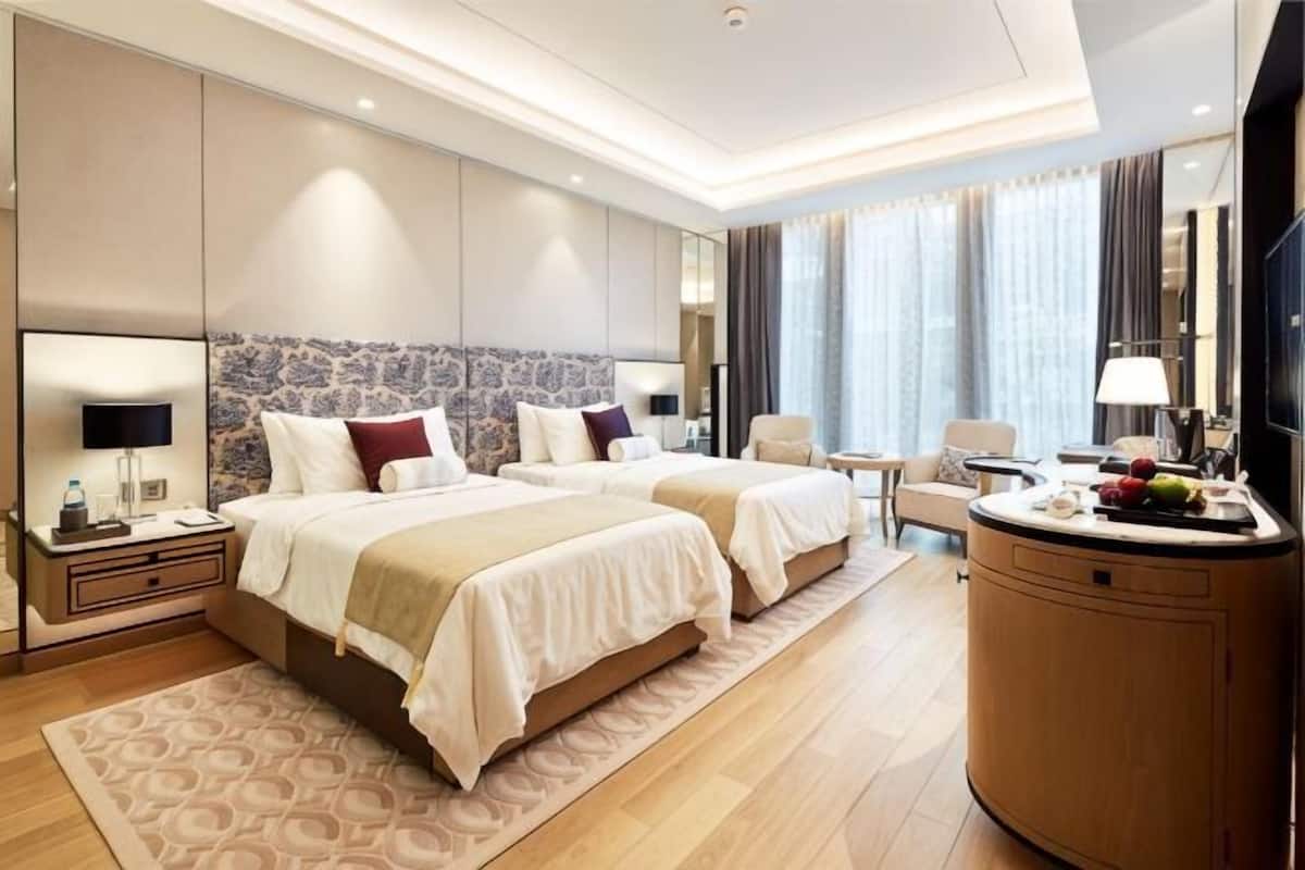 Quarto de hotel moderno com duas camas, uma penteadeira redonda de madeira, uma escrivaninha e um espelho grande.