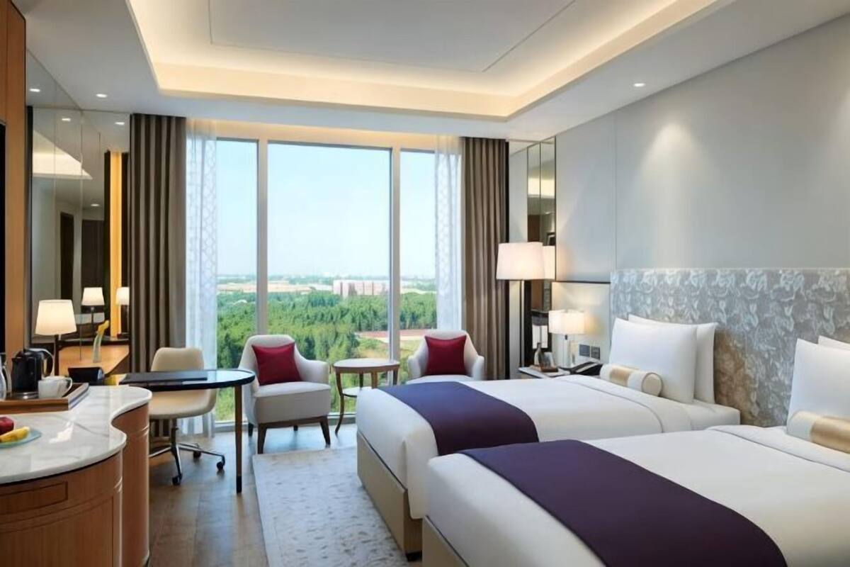 Quarto de hotel com duas camas, uma escrivaninha, uma cadeira, uma mesinha e vista para a cidade.