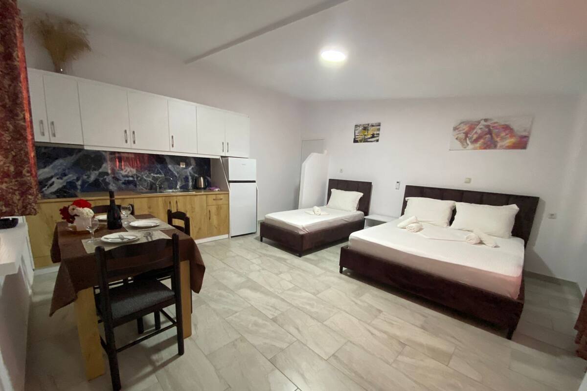 A compact hotel room with a kitchenette, two single beds, a dining table, and a large window.