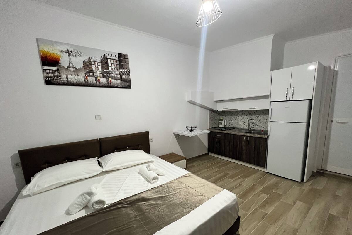 A hotel room with a bed, a small kitchenette, a white refrigerator, and a painting on the wall.