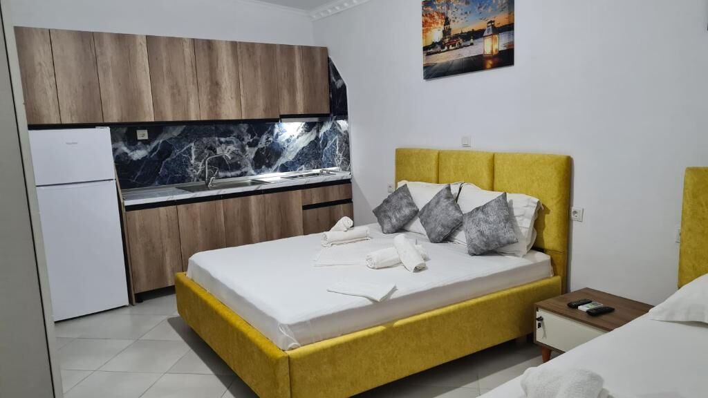 A bedroom with a bed, a nightstand, a small table, a kitchen area with a marble countertop, and wooden cabinets.