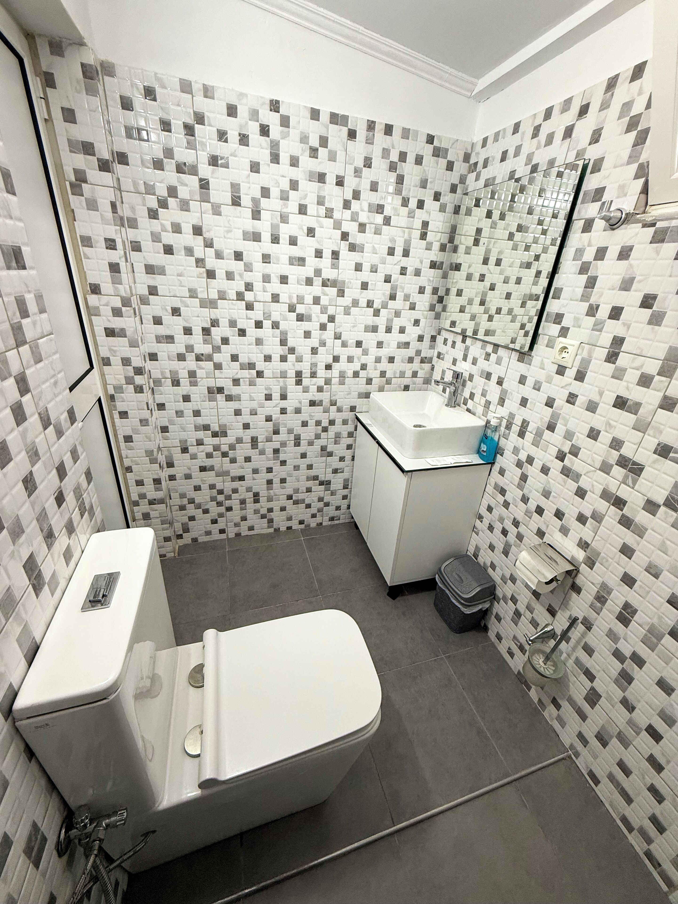 A bathroom with a toilet, sink, and mirror. The walls are tiled with a checkered pattern.