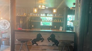 A bar area with wooden flooring, two stools, hanging pendant lights, and a counter with shelves displaying various items. - Kings Court (Dalton)