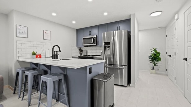 A modern kitchen with grey cabinets, a stainless steel refrigerator, a microwave, and a bar stool area. - King Bed Mtn Escape Free Parking 1hr to Banff (Calgary)