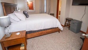 Quarto com cama, mesa de cabeceira, luminária e TV fixada na parede.