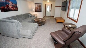 Uma sala de estar com um sofá seccional cinza, uma poltrona de couro marrom, duas mesas laterais de madeira e uma TV de tela plana na parede.
