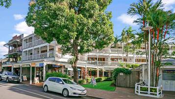 A street view with a white car parked on the side, a two-story building with balconies, and a palm tree.