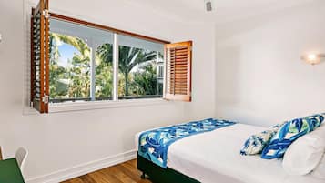 A bedroom with a bed, a window with shutters, a ceiling fan, and a view of palm trees outside.