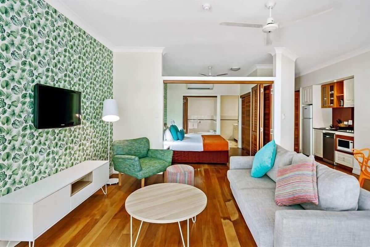 Una habitación de hotel moderna con televisión de pantalla plana, un sofá, una mesa de comedor, una cama con cabecera de madera y una zona de cocina.