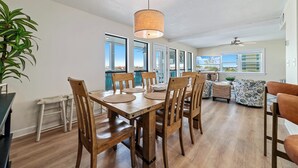 Un vaste espace dédié à la salle à manger, avec une table et des chaises en bois, un luminaire suspendu et de grandes fenêtres offrant une vue sur l’extérieur.