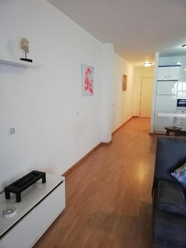 Una sala de estar con sofá, una mesa de centro y un mueble para la televisión. Se ve un pasillo que conduce a una zona de cocina.