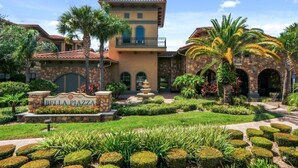 A multi-story building with a stone facade, a sign reading "Bella Piazza," and a landscaped garden with palm trees and trimmed hedges.