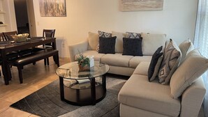 A living room with a sectional sofa, a glass coffee table, a dining table with chairs, and a chandelier.