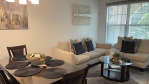 A modern living room with a dining area, featuring a sofa set, a glass coffee table, and a dining table with placemats.