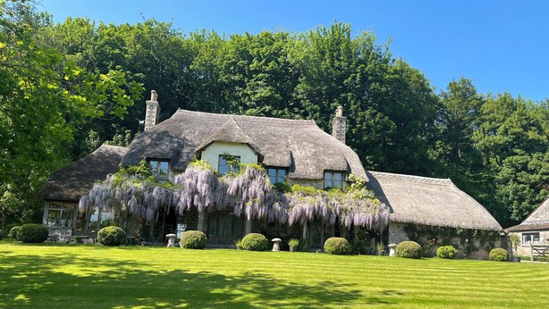 Une maison traditionnelle en pierre, avec un toit de chaume, entourée d’une végétation luxuriante et d’une pelouse bien entretenue.