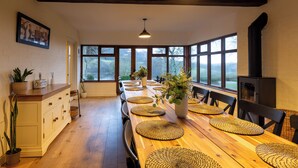 Un coin repas avec une longue table en bois, agrémentée de sets de table, de chaises et d’un vase central orné de fleurs. Un meuble avec des tiroirs et une plante en pot se trouvent sur la gauche.