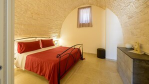 Un dormitorio con techo de piedra, una cama con ropa de cama roja, una ventana pequeña con cortinas y un aparador de madera con un reloj.