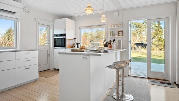 A modern kitchen with white cabinetry, a central island with a sink, a built-in oven, and a large window providing a view of the outdoors. - 3 bedroom awesome home in Odder (Odder)