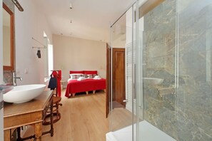 Una habitación de hotel moderna con una cama grande, un escritorio de madera, un ventanal amplio y un baño con lavamanos blanco.