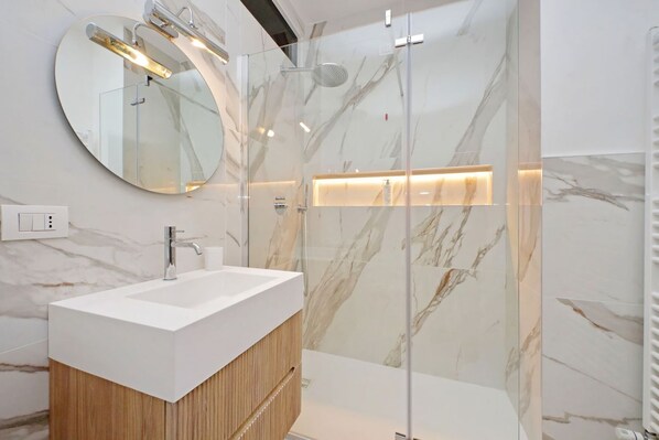 Une salle de bain moderne avec un lavabo blanc, un meuble-lavabo en bois, une cabine de douche en verre et des murs en marbre.
