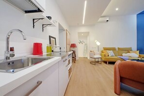 Una cocina moderna con un lavamanos, armarios y una tetera roja. En la zona de estar, hay un sofá amarillo y una silla blanca.