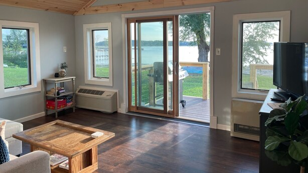Ein geräumiges Zimmer mit einer großen Glasschiebetür, die auf eine Terrasse führt, Holzbalken an der Decke, einem Deckenventilator und einem Fernseher auf einem Schrank.
