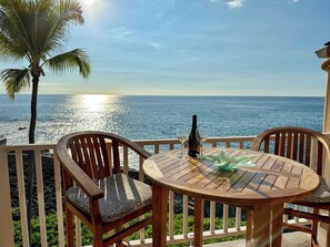 Een houten tafel met twee stoelen en een fles wijn, met uitzicht op een strand met palmbomen en de oceaan.