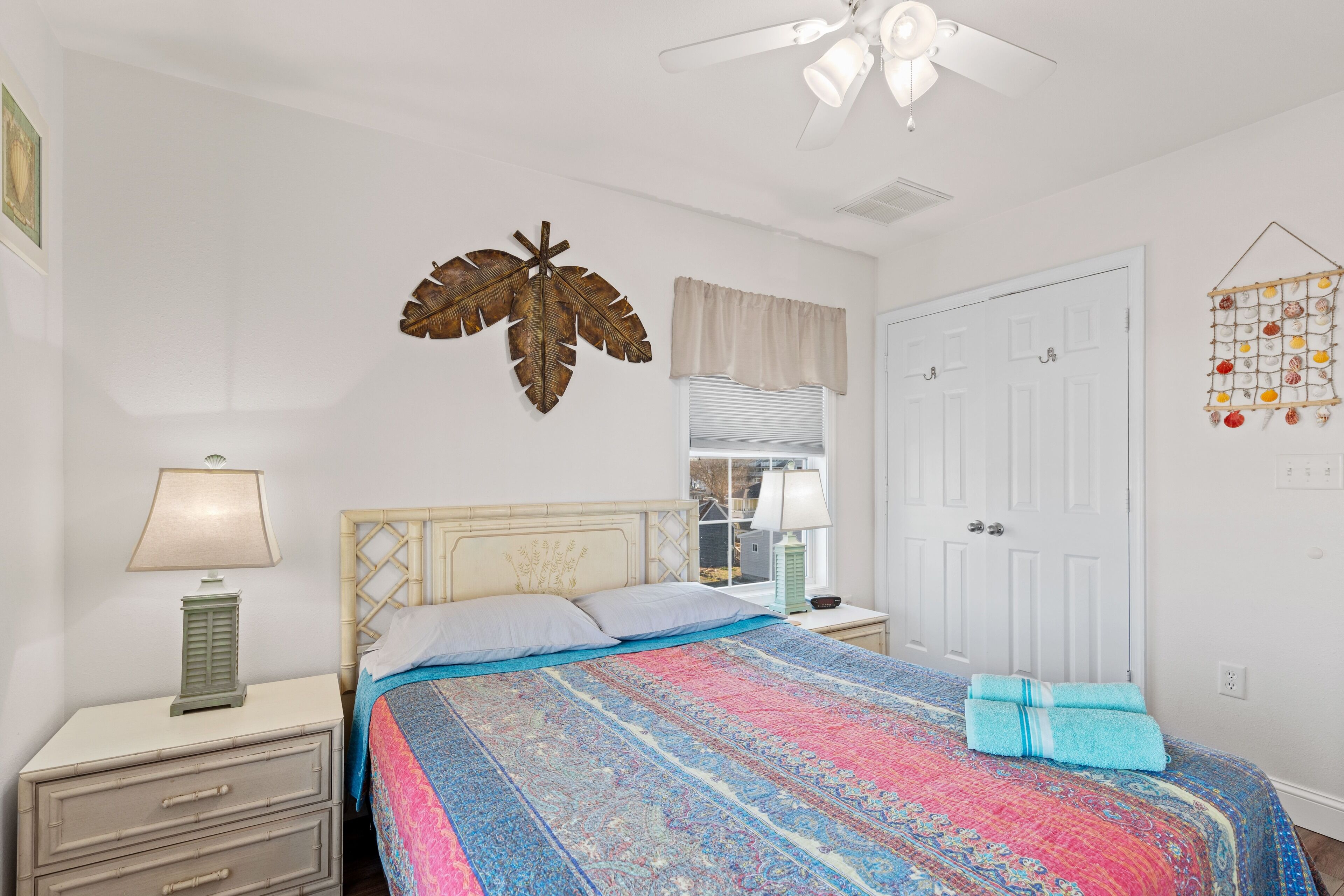 A bedroom with a bed, nightstands, a ceiling fan, a window with blinds, and a wall decoration.